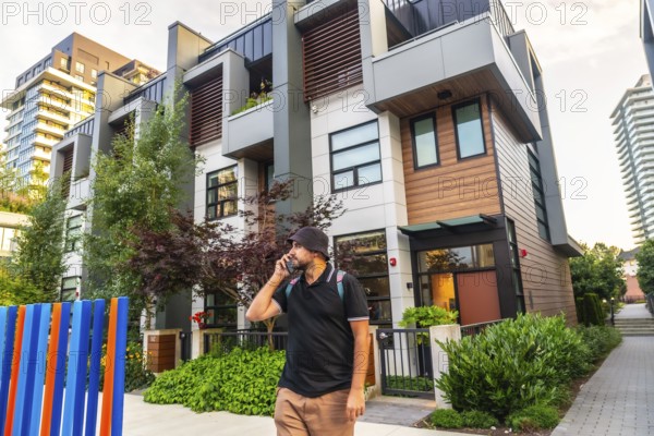 Young man with backpack is using his phone while walking in front of modern townhouses in vancouver, british columbia, canada, showcasing urban lifestyle and real estate