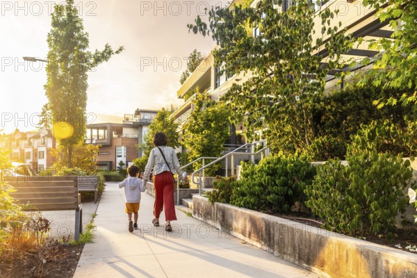 Mother and son holding hands are walking away from the camera on a sidewalk in a residential neighborhood of vancouver, with warm sunset light illuminating the scene
