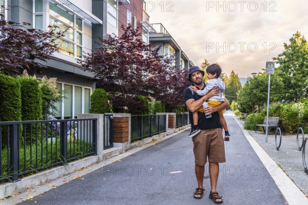 Father carrying his son while walking on a sidewalk in a residential area of vancouver, british columbia, with modern townhouses, trees, and a bus stop sign in the background during a sunny day