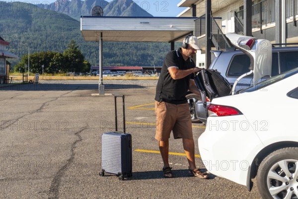 Tourist wearing headphones is loading luggage into car trunk in motel parking lot, preparing for road trip adventure in scenic mountain landscape
