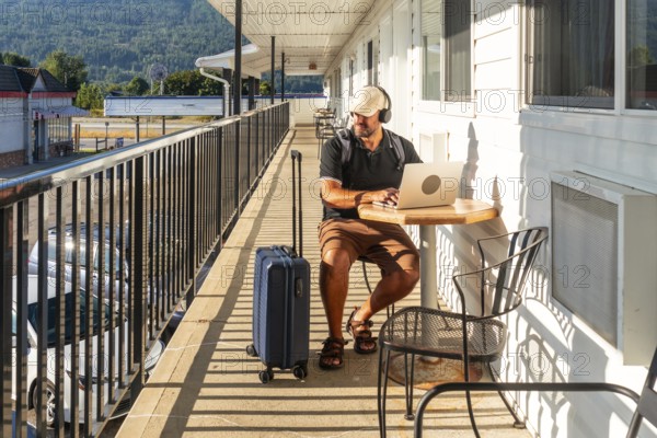 Male digital nomad wearing headphones and working on laptop while sitting at table on motel balcony with luggage nearby, enjoying sunny morning and mountain view