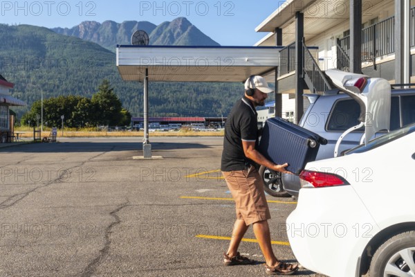 Tourist wearing headphones is loading a suitcase into the trunk of his car in the parking lot of a motel, ready to start his road trip adventure in the mountains