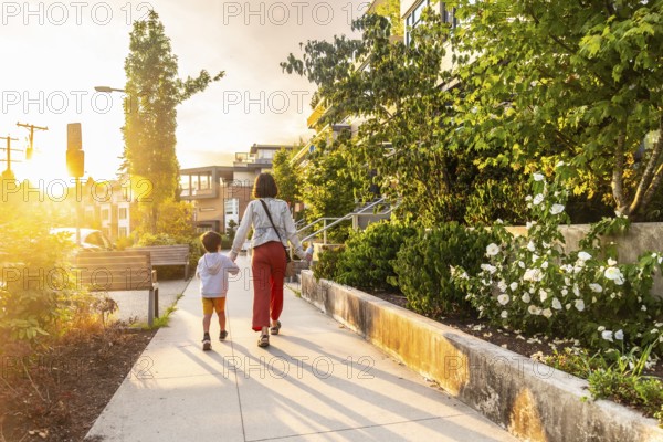 Mother and son are walking hand in hand down a sidewalk in a residential neighborhood of vancouver, british columbia, during a beautiful sunset, enjoying quality time together