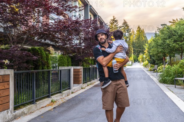 Father carrying his son is walking down a residential street in vancouver, british columbia, enjoying the pleasant summer evening and the urban landscape surrounding them