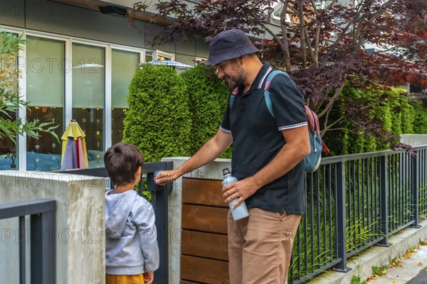 Father holding a reusable water bottle and wearing a bucket hat opens the gate for his son at a daycare in vancouver, british columbia, starting their morning drop off routine