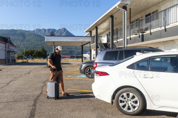 Male tourist wearing headphones and carrying a backpack strolls through a parking lot, pulling a rolling suitcase toward a motel after arriving by car for a summer getaway