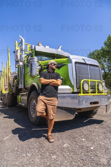 Truck driver confidently posing with arms crossed, leaning against a modern green logging truck in kamloops, british columbia, under a bright sunny sky on a warm summer day