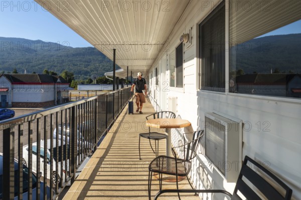 Tourist walking along a motel balcony, rolling luggage in tow, passing by a table and chairs, enjoying a sunny morning with majestic mountains providing a stunning backdrop