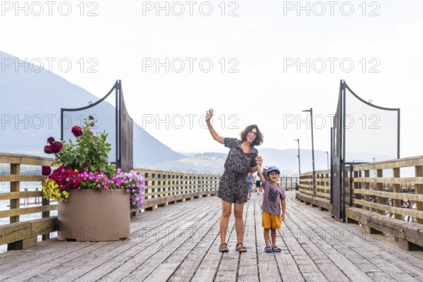Cheerful mother and her young son are waving while standing on a picturesque wooden pier in salmon arm, british columbia, enjoying the scenic beauty of the shuswap lake and surrounding mountains