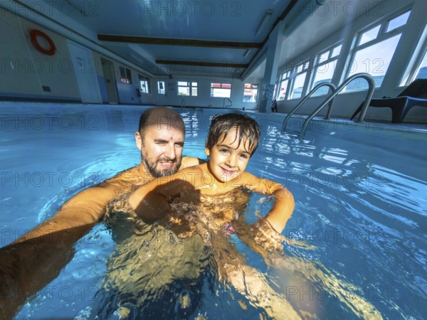 Father and son enjoying a playful moment while taking a selfie in an indoor swimming pool, creating lasting memories during their leisure time together