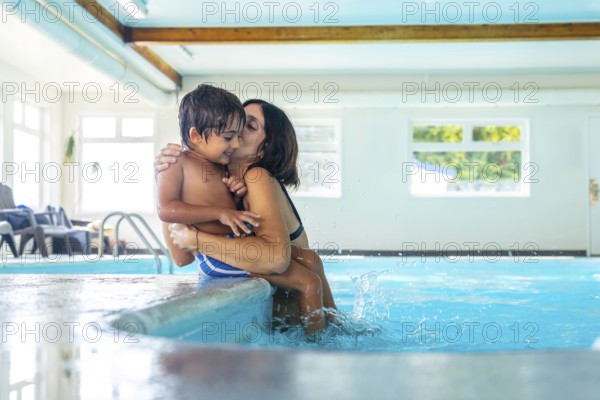 Mother holding and kissing her son at the poolside, with his feet splashing in the clear water, creating playful ripples and capturing moments of joy and affection