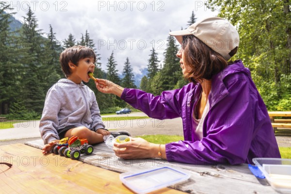 Mother feeds her son a healthy snack at a picnic table in hemlock grove, a beautiful boardwalk in the canadian rockies, surrounded by lush green trees and a tranquil atmosphere