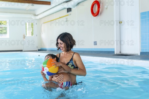 Mother holding her son while playing with a vibrant inflatable beach ball in an indoor swimming pool, creating joyful family memories during a fun filled leisure moment together