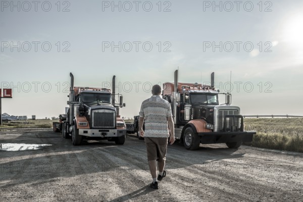 Trucker walking towards two semi trucks parked on a gravel road near hemlock grove boardwalk in the canadian rockies, showcasing a scene of transportation and logistics