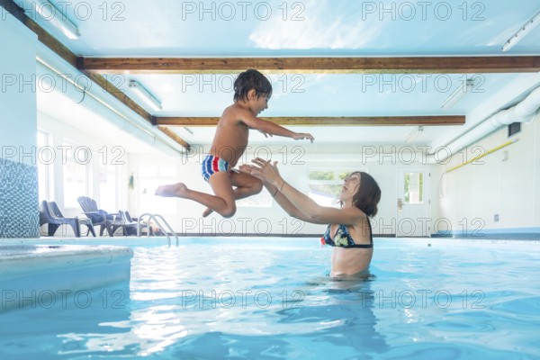 Smiling young mother catching her cheerful son as he leaps joyfully into the indoor swimming pool, creating a moment of fun and bonding during their summer vacation