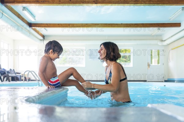 Happy mother and son enjoying playful moments at the edge of an indoor swimming pool, splashing water and sharing laughter during a fun filled leisure activity, creating joyful memories together