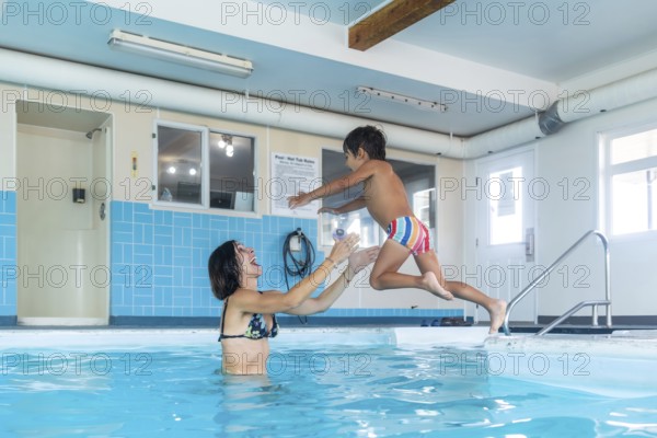 Happy mother catching her smiling son mid jump into a swimming pool at a lively public indoor aquatic center, celebrating joy, fun, and togetherness in a vibrant family moment