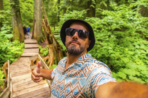 Happy tourist wearing sunglasses and bucket hat taking a selfie while hiking the beautiful giant cedars boardwalk trail in mount revelstoke national park, british columbia, canada, giving thumbs up