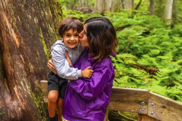 Mother kissing her smiling son while hiking the giant cedars boardwalk trail in mount revelstoke national park, british columbia, canada, surrounded by lush green ferns