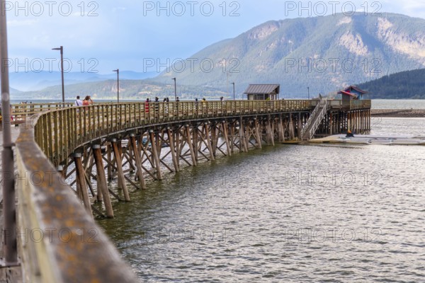 Tourists are walking on the long wooden pier that curves across the shuswap lake in salmon arm, british columbia, with mountains and cloudy sky in the background on a sunny summer day