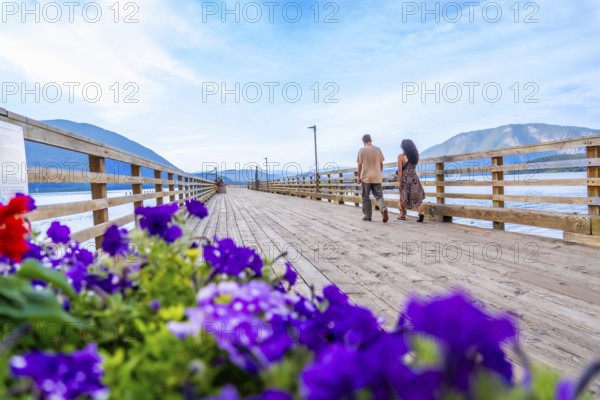 Tourists walking on a wooden pier adorned with vibrant flowers, enjoying the scenic beauty of salmon arm in british columbia, canada, with mountains and blue sky in the background