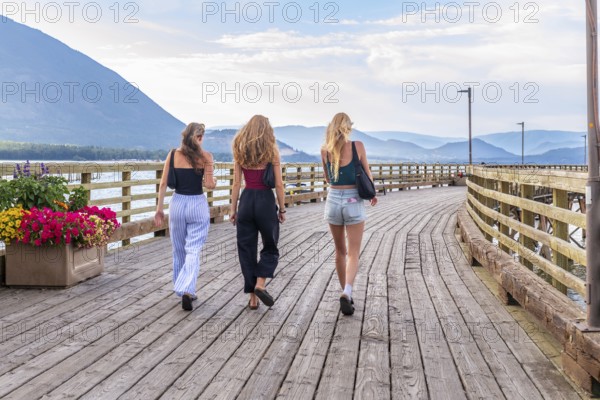 Three young women are enjoying a leisurely stroll along a wooden pier in salmon arm, british columbia, taking in the scenic beauty of the shuswap lake and surrounding mountains