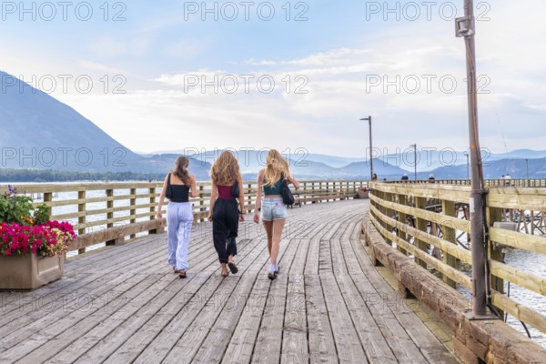 Three young women are enjoying a leisurely stroll along a scenic wooden pier in salmon arm, british columbia, surrounded by breathtaking views of the shuswap lake and surrounding mountains