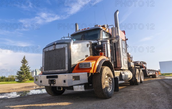 Gleaming orange and silver semi truck parked on a gravel road, bathed in the warm glow of late afternoon sunlight, against a backdrop of a clear blue sky and a serene rural landscape