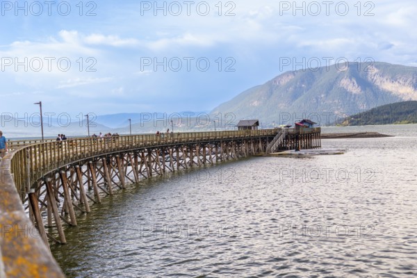Tourists are enjoying a sunny day walking on the longest wooden wharf in north america, located in the city of salmon arm, british columbia, canada, with a mountain range in the background