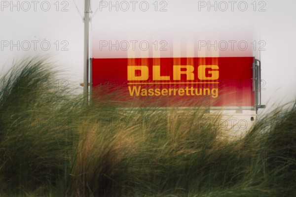 DLRG sign on the coast, red sign with the inscription DLRG Wasserrettung partly covered by high dune grass, windy scenery with moving grass and dimmed daylight (ICM), Schillig, Lower Saxony, Germany