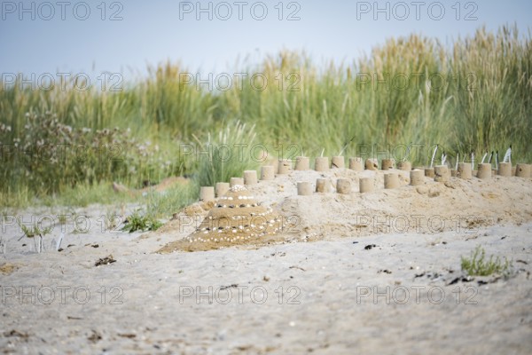 Sandcastle on the beach, small sandcastle in the foreground in front of overgrown dunes with beach grass, bright daylight under blue sky and quiet, summery coastal atmosphere, Schillig, Lower Saxony, Germany
