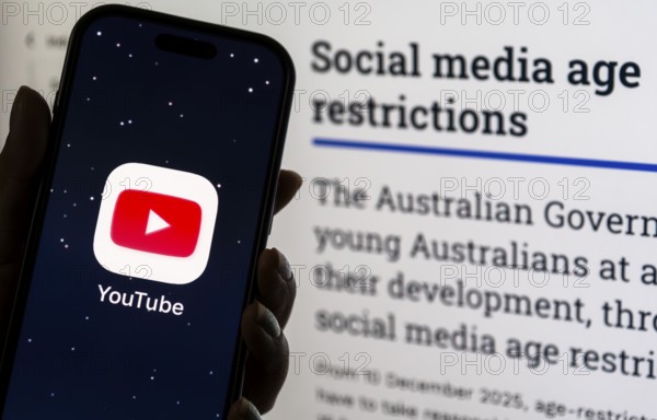 In this photo illustration, a person holds a phone displaying the YouTube app in front of a screen showing information about Australia's new social media age restrictions, in New Delhi, India on 12 December 2025