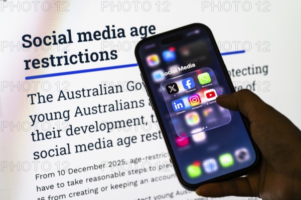 In this photo illustration, a person holds a smartphone displaying popular social media apps in front of a screen showing information about Australia's new social media age restrictions, in New Delhi, India on 12 December 2025
