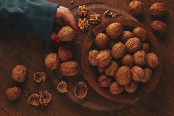 A woman's hand taking a walnut from a wooden bowl filled with walnuts. There are shells and walnuts scattered around