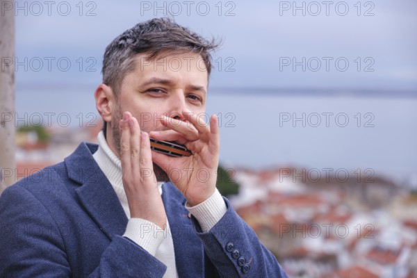 Man musician in a blue blazer and white sweater playing blues on a harmonica with eyes closed, standing outdoors in city street