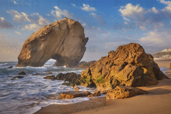 Penedo do Guincho, a large boulder rock arch at Praia da Santa Cruz, Portugal, with ocean waves and sandy beach on sunset