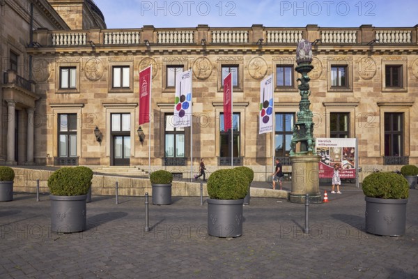 Rheingau Music Festival, spa hotel Wiesbaden, advertising flags on flagpoles, spa hotel, building material sandstone, architectural style neoclassicism, facade with windows, planter with boxwood (Buxus sempervirens), bollards, blue sky, evening light, sunlight, Kurhausplatz, Wiesbaden, state capital, independent city, Hesse, Germany