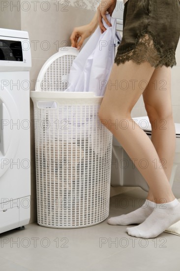 Unrecognizable woman stands next to a washing machine. She is putting white clothes into a laundry basket. The room has tile flooring and a washing machine nearby