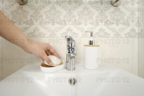 A hand reaches for a bar of soap placed on a dish near a sink. Water flows from a silver faucet into the basin. A soap dispenser sits beside the sink in a bathroom setting