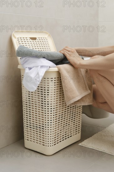 A person is sorting towels and clothes into a laundry basket in a bathroom. The setting is simple, with a tiled wall and a light colored basket. The person is bent over the basket