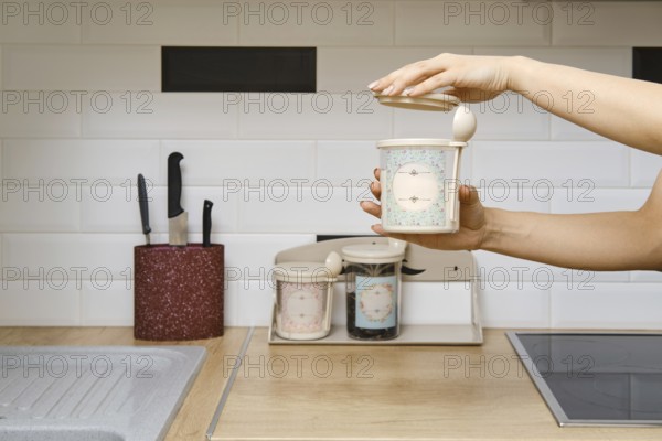 A person holds a container in one hand while opening the lid with the other. The kitchen has a clean counter with storage jars and knives visible nearby. The setting is bright and well-organized