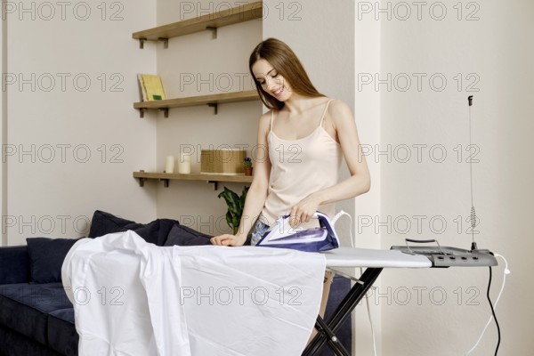 A woman stands next to an ironing board in a bright living room. She presses a white shirt with an iron. The room has shelves with books and decorations