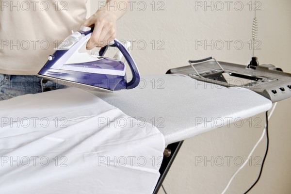 A person stands by an ironing board and uses an iron to press a shirt. The setting is a home environment during the afternoon. The task focuses on removing wrinkles from clothing