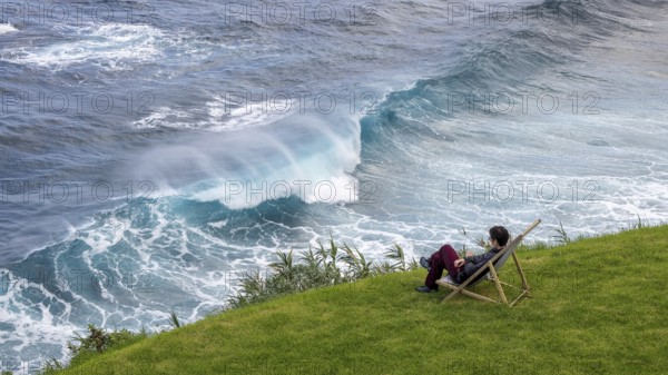 Woman sitting comfortably in deckchair in a meadow and watching waves in the sea, Madeira, Portugal