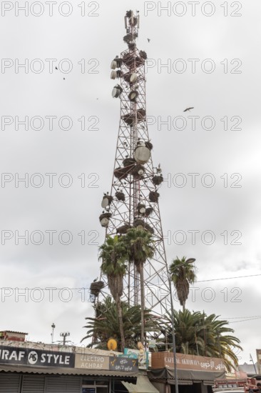 Tall radio tower with satellite dishes and palm trees in an urban setting under cloudy skies in Morocco