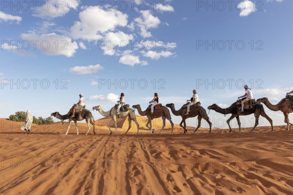 Caravan of camels with riders crossing the sandy desert under cloudy sky, Sahara, Morocco
