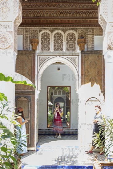 A traditional courtyard with arches, mirrors and decoration and two woman, Bahia Palace in Marrakech, Morocco