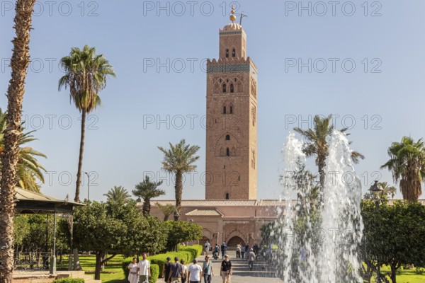 The Koutoubia Mosque in Marrakech with palm trees and a fountain in a sunny garden, visited by tourists