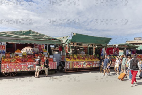 Market stalls full of fruit with vendors and passers-by on a sunny day