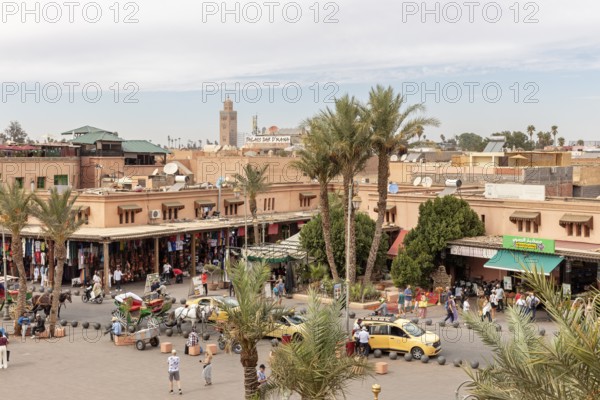 Bustling market scene with palm trees and buildings, a minaret and a blue sky in the background